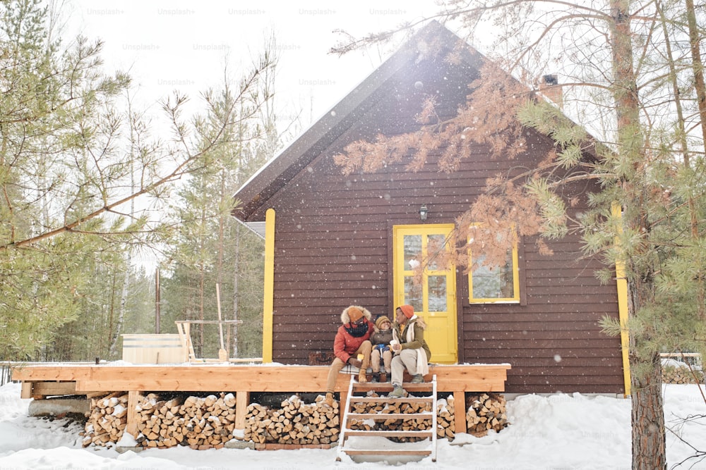 冬服を着た3人の幸せな家族が、カントリーハウスやコテージのそばの木製ポーチでおしゃべりをしながら、そこで休暇を過ごす