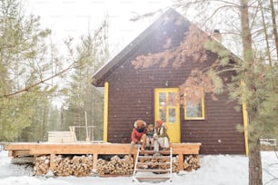 Glückliche dreiköpfige Familie in Winterkleidung, die sich auf der Holzveranda bei ihrem Landhaus oder Ferienhaus unterhält, während sie dort Urlaub macht