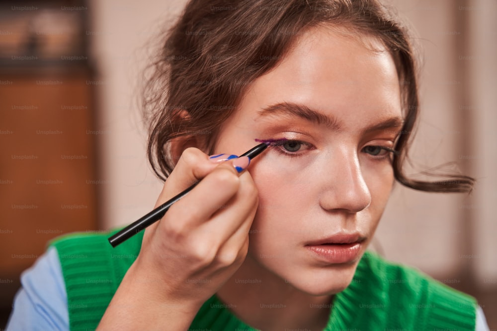 Close up view of the teenage girl holding brush with eyelids and looking attentively at the mirror while making make up during the weekend