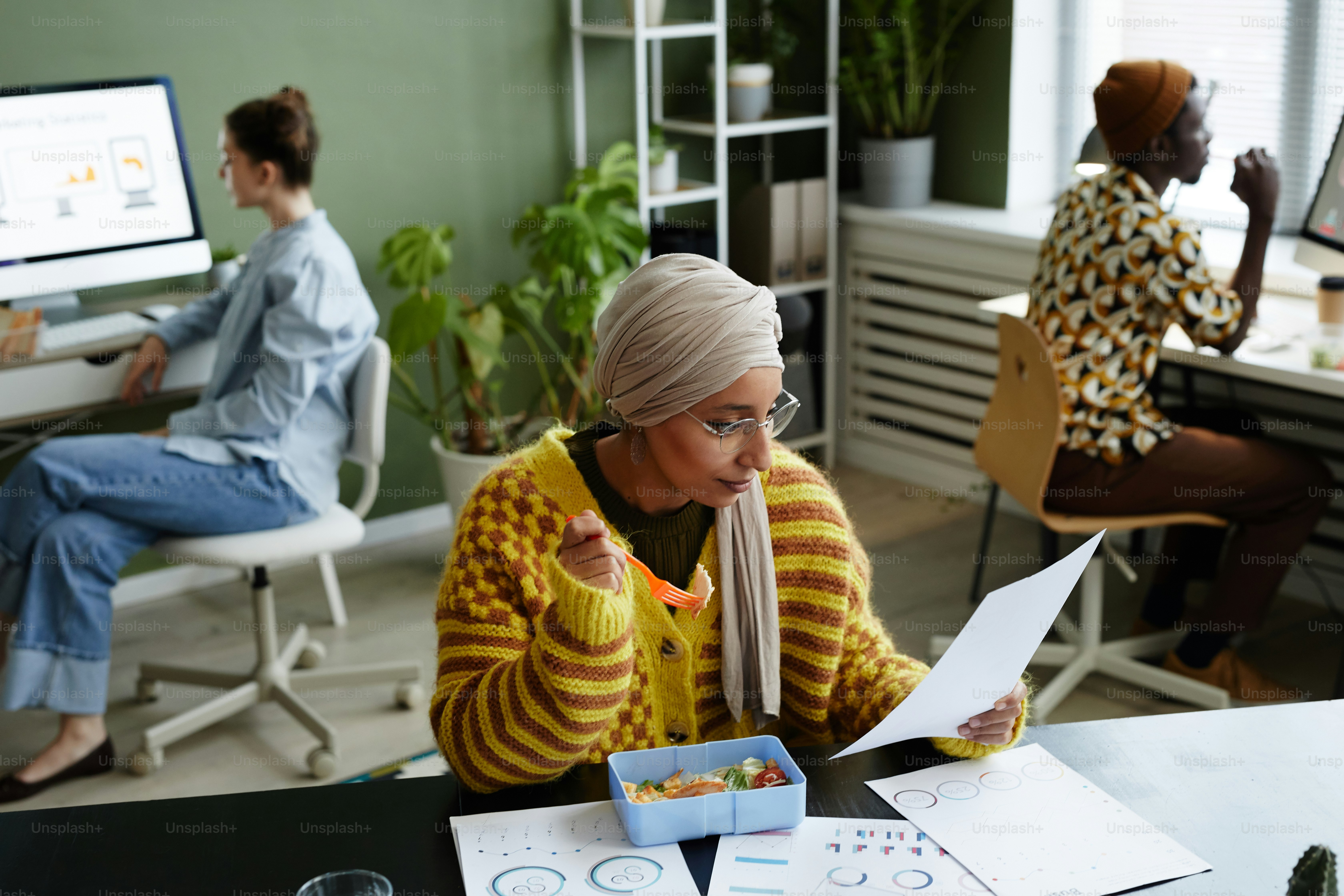Portrait of ethnic young woman wearing headscarf and eating healthy lunch in office during work