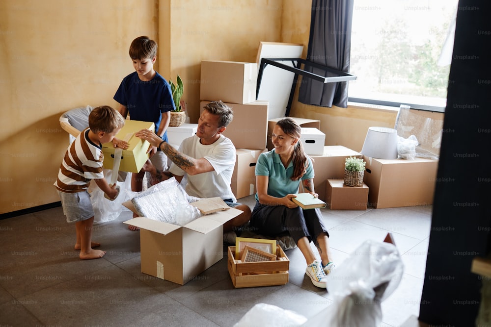 Ganzkörperporträt einer glücklichen Familie mit zwei Kindern, die beim Umzug gemeinsam Kisten auspacken, Kopierraum