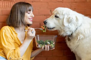 Young woman feeding her cute dog, while sitting together and eating healthy salad indoors. Friendship and spending time with pets concept