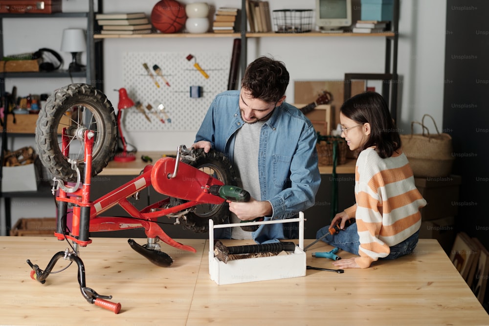 전기 드릴을 사용하는 젊은 남자는 딸이 근처에 앉아 아버지를 돕는 동안 나무 테이블로 자전거 바퀴를 수리한다