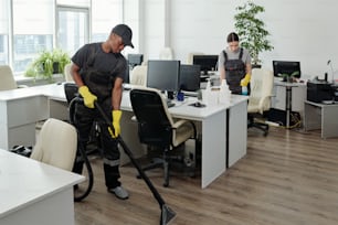 Dos jóvenes trabajadores de una empresa de servicios de limpieza contemporánea en overoles y guantes que realizan su trabajo en una oficina de espacio abierto