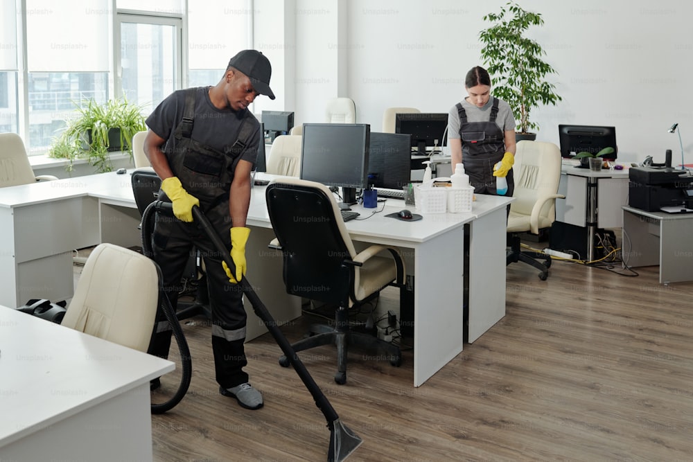 Two young workers of contemporary cleaning service company in coveralls and gloves carrying out their work in openspace office