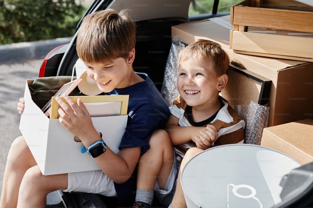 Ritratto di due ragazzi sorridenti seduti nel bagagliaio dell'auto con scatole mentre si trasferiscono nella nuova casa