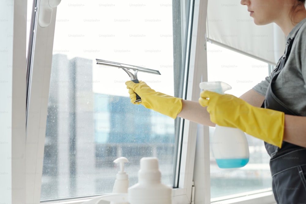 Manos de una joven limpiadora con guantes amarillos y ropa de trabajo lavando grandes ventanas de oficina con detergente y espátula de silicona