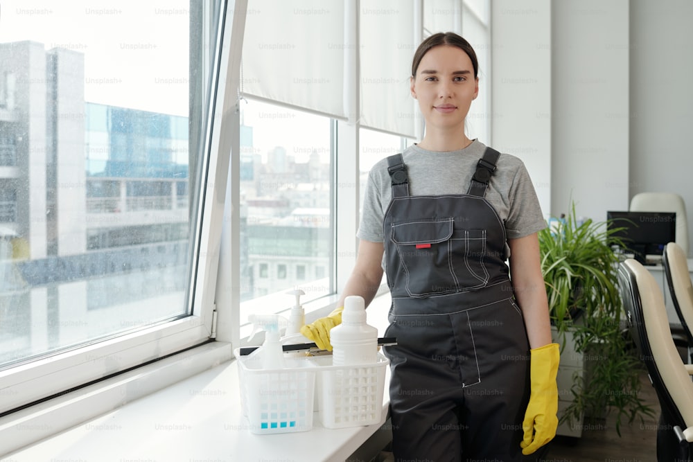 Personal femenino joven de una empresa de limpieza contemporánea de pie junto a la ventana en una gran oficina de espacio abierto y mirando a la cámara