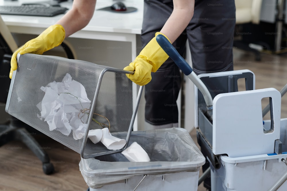 Mãos enluvadas de faxineira jogando lixo da lixeira em balde plástico no carrinho de zelador enquanto trabalhava no escritório moderno