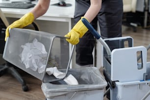 Manos enguantadas de una limpiadora tirando basura del cubo de basura en un cubo de plástico en el carrito de conserje mientras trabajaba en una oficina moderna