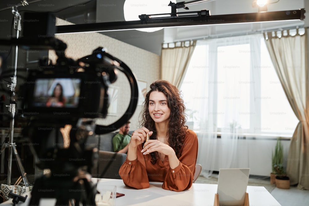 Happy young female making speech in front of camera during commercial record in studio or living room of loft apartment