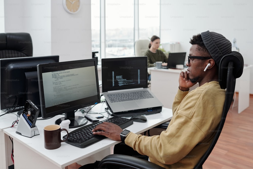 Seitenansicht eines jungen, ernsthaften Mannes, der sich auf die Dekodierung von Daten konzentriert, während er vor dem Computermonitor am Arbeitsplatz im Büro sitzt