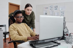 Two contemporary programmers decoding data on laptop screen by workplace while young African American man typing on keyboard