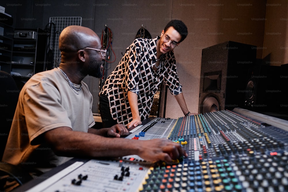 Productor musical afroamericano maduro y joven cantante charlando sobre algo divertido mientras trabaja en un estudio de grabación