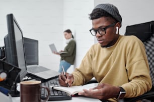Young serious black man in casualwear making notes by workplace while sitting against female coworker with laptop standing by wall