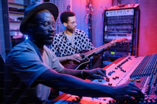 스타일리시한 성숙한 아프리카계 미국인 프로듀서가 믹서에서 소리를 조절하는 동안 젊은 음악가가 기타를 연주하는 장면