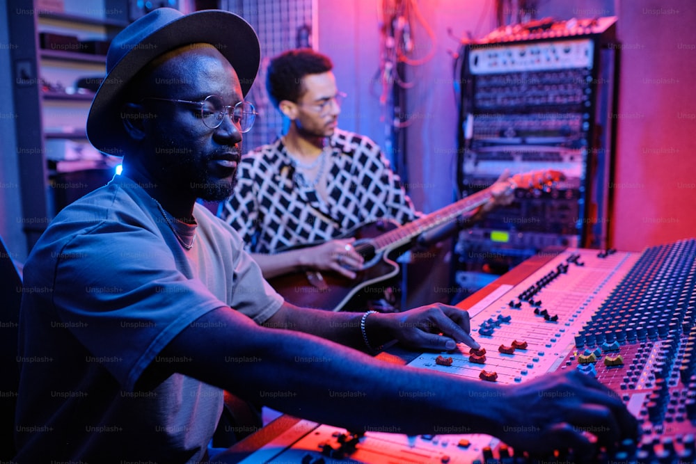 Selective focus shot of stylish mature African American producer adjusting sound on mixer while young musician playing guitar