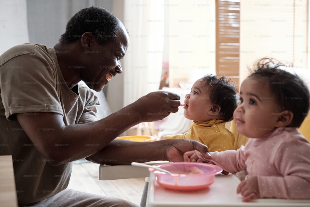 Cheerful mature African American father taking care of his baby daughters feeding them with fruit puree