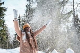 Verspielte junge Afroamerikanerin mit langen Haaren, die Spaß daran hat, an einem sonnigen Wintertag Schnee in die Luft zu werfen