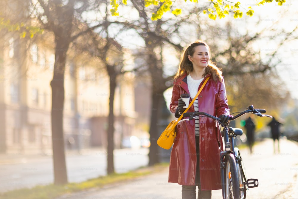 Lächelnde stilvolle Frau mittleren Alters im roten Regenmantel mit Fahrrad und Tasse Cappuccino, die draußen auf der Stadtstraße spazieren geht.