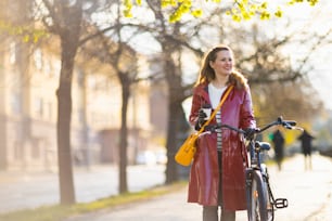 smiling stylish middle aged woman in red rain coat with bicycle and cup of cappuccino walking outside on the city street.