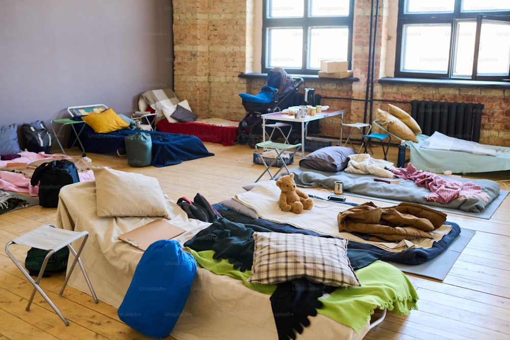 Gran grupo de lugares para dormir con mantas, almohadas, juguetes, sacos y otros elementos esenciales preparados para los refugiados en una habitación espaciosa