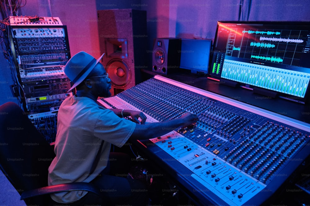 帽子をかぶったスタイリッシュな成熟したアフリカ系アメリカ人の男性が、ネオンライトの中でレコーディングスタジオでミキシングコンソールを使用してサウンドトラックを作成する