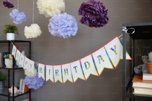 Imagen de banner con texto de color Feliz cumpleaños colgado en dos estantes con flores de papel encima decorando para la fiesta de cumpleaños