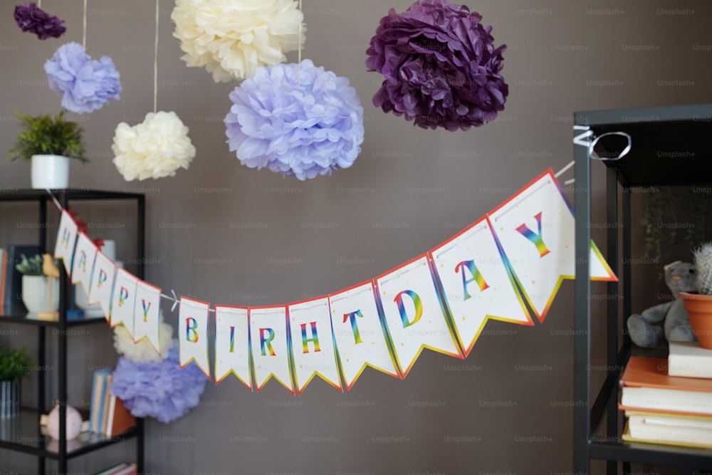 Bild eines Banners mit farbigem Text Happy Birthday, das auf zwei Regalen mit Papierblumen darüber hängt, die für die Geburtstagsfeier dekoriert sind