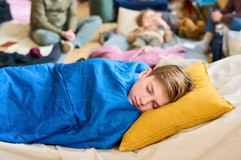 Peaceful schoolboy napping on sleeping place under blue blanket while keeping his head on yellow pillow against group of refugees
