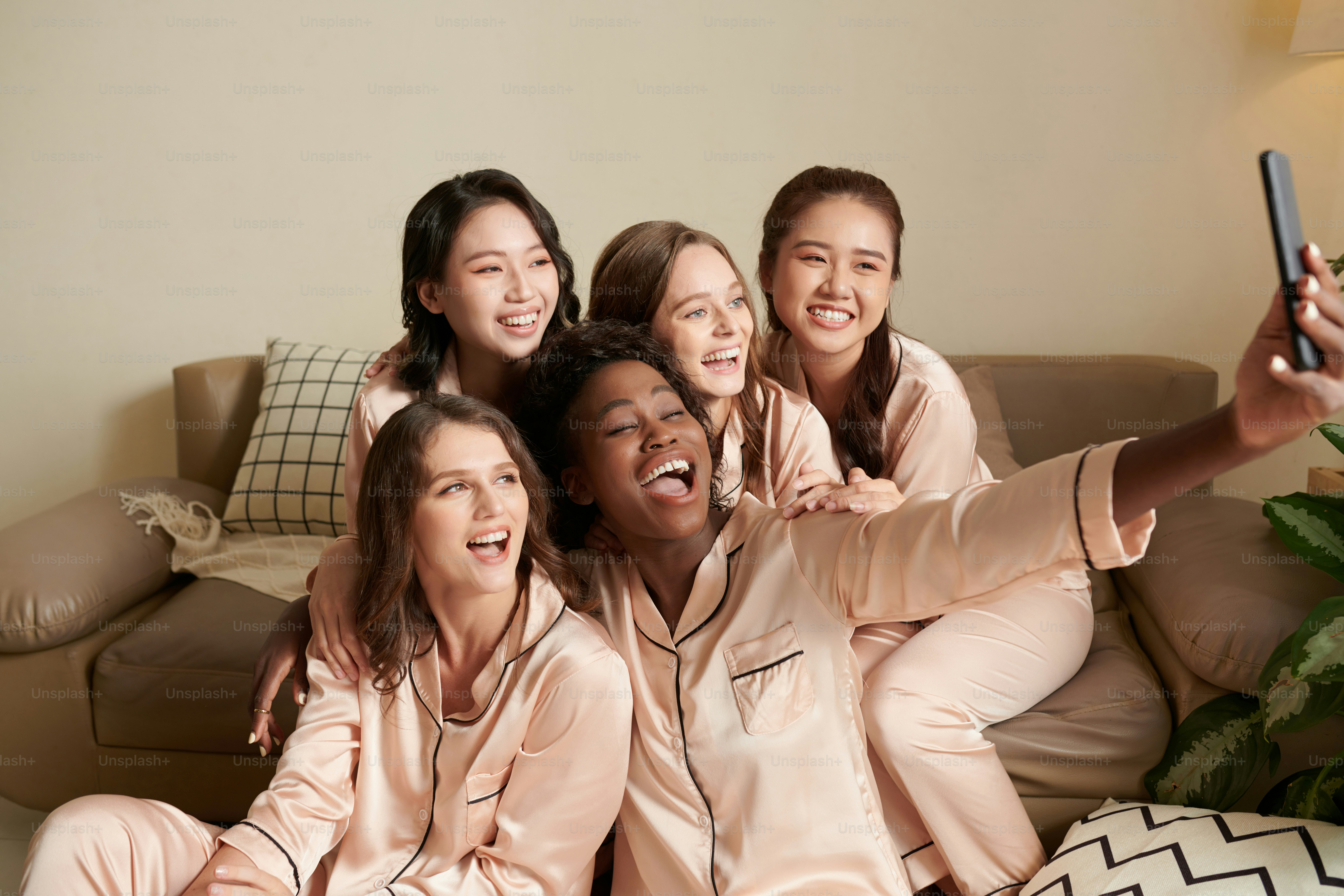Diverse group of smiling young women in silk pajamas taking selfie ...