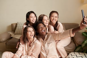 Joyful female friends in satin pajams taking selfie on smartphone