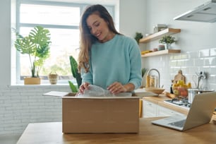 Attractive young woman unpacking box while standing at the domestic kitchen