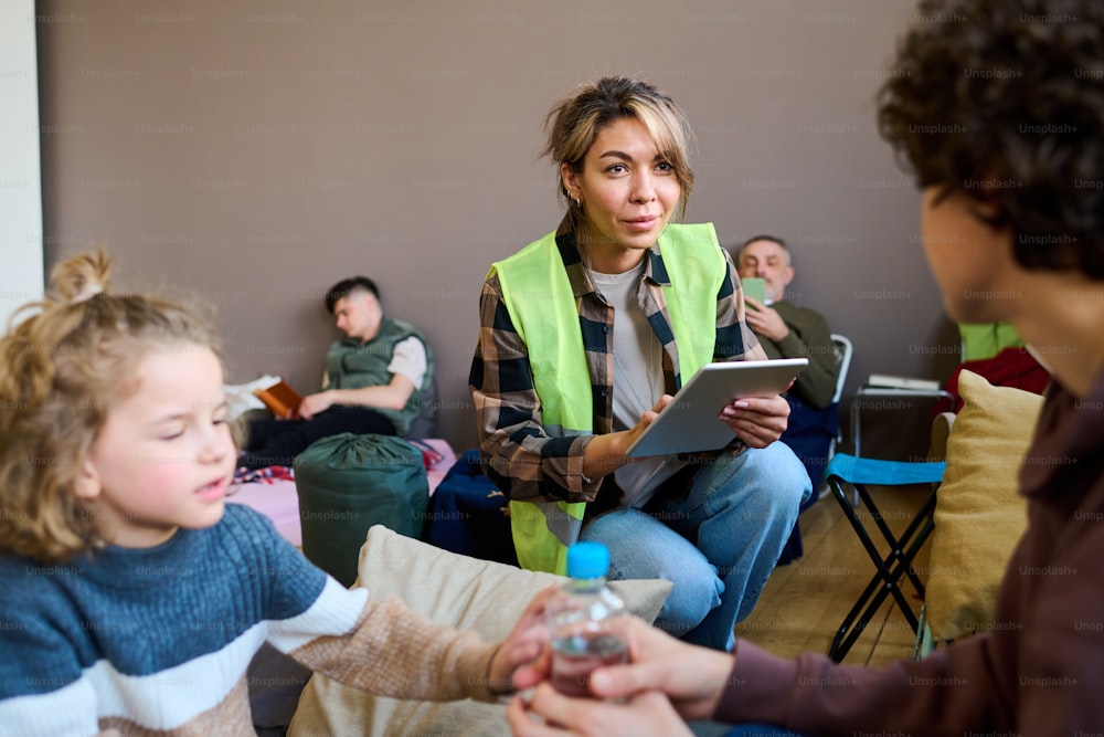 Jeune bénévole avec tablette assise sur des squats devant une femme avec son fils et entrant leurs noms et autres données personnelles