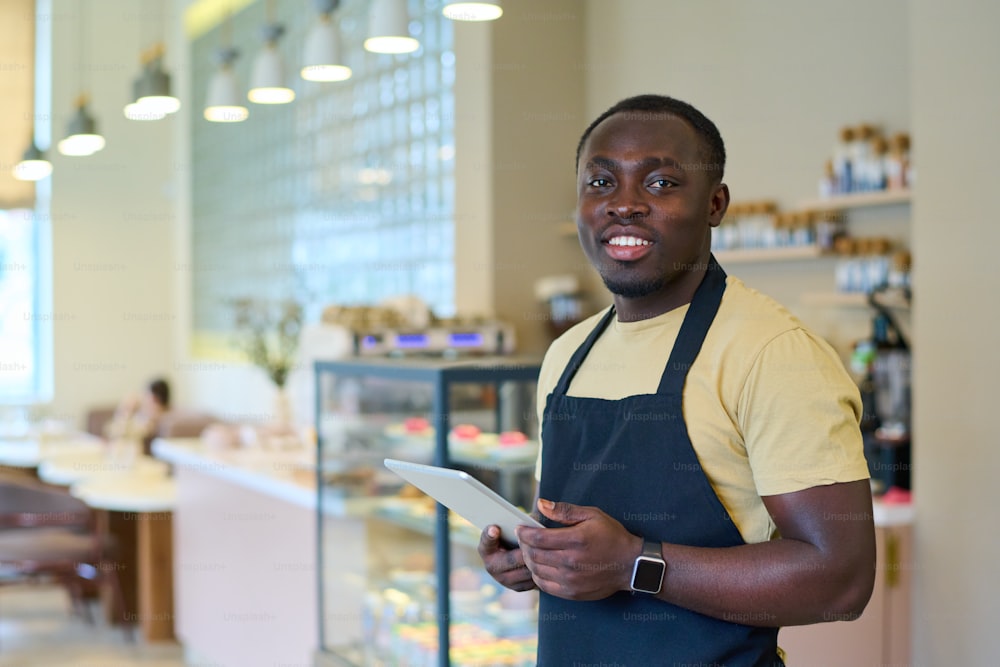 Portrait of African young waiter in uniform smiling at camera while taking order online on tablet pc