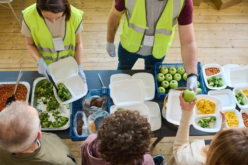 Acima do ângulo de dois voluntários uniformizados espalhando comida entre refugiados com contêineres em fila por mesa