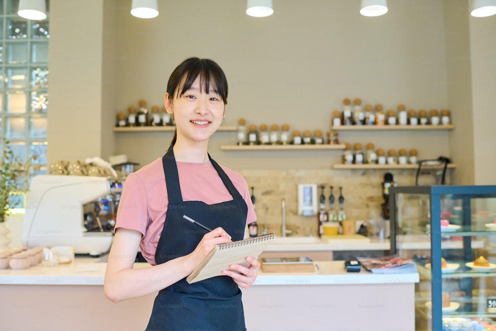 Portrait of Asian young waitress in apron smiling at camera and making notes in notepad while standing in coffee shop