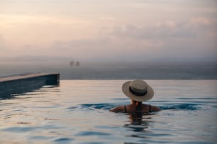 インフィニティプールでリラックスし、美しい夕日と海の景色を眺める帽子をかぶった女性の背面図