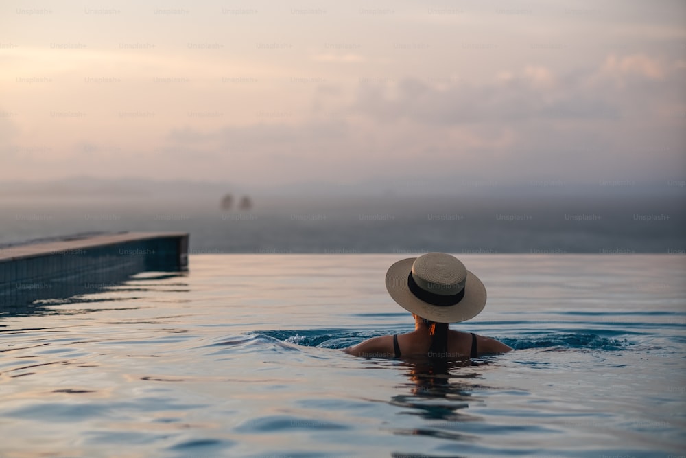 Rückansicht einer Frau mit Hut, die sich im Infinity-Pool entspannt und einen wunderschönen Sonnenuntergang und den Meerblick betrachtet