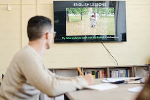 Enfoque selectivo en la toma de fondo de un joven irreconocible sentado en la mesa viendo un video o una presentación durante la clase de inglés
