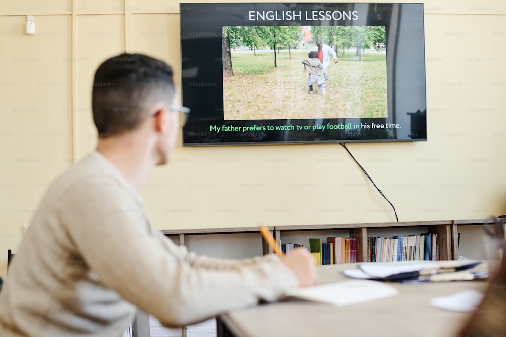 Selective focus on background shot of unrecognizable young man sitting at table watching video or presentation during English lesson