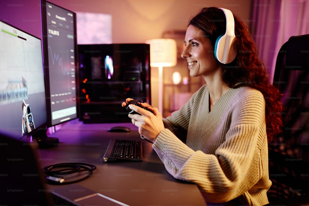 Mujer caucásica adulta joven moderna con auriculares jugando al videojuego de disparos usando el controlador de la consola por la noche