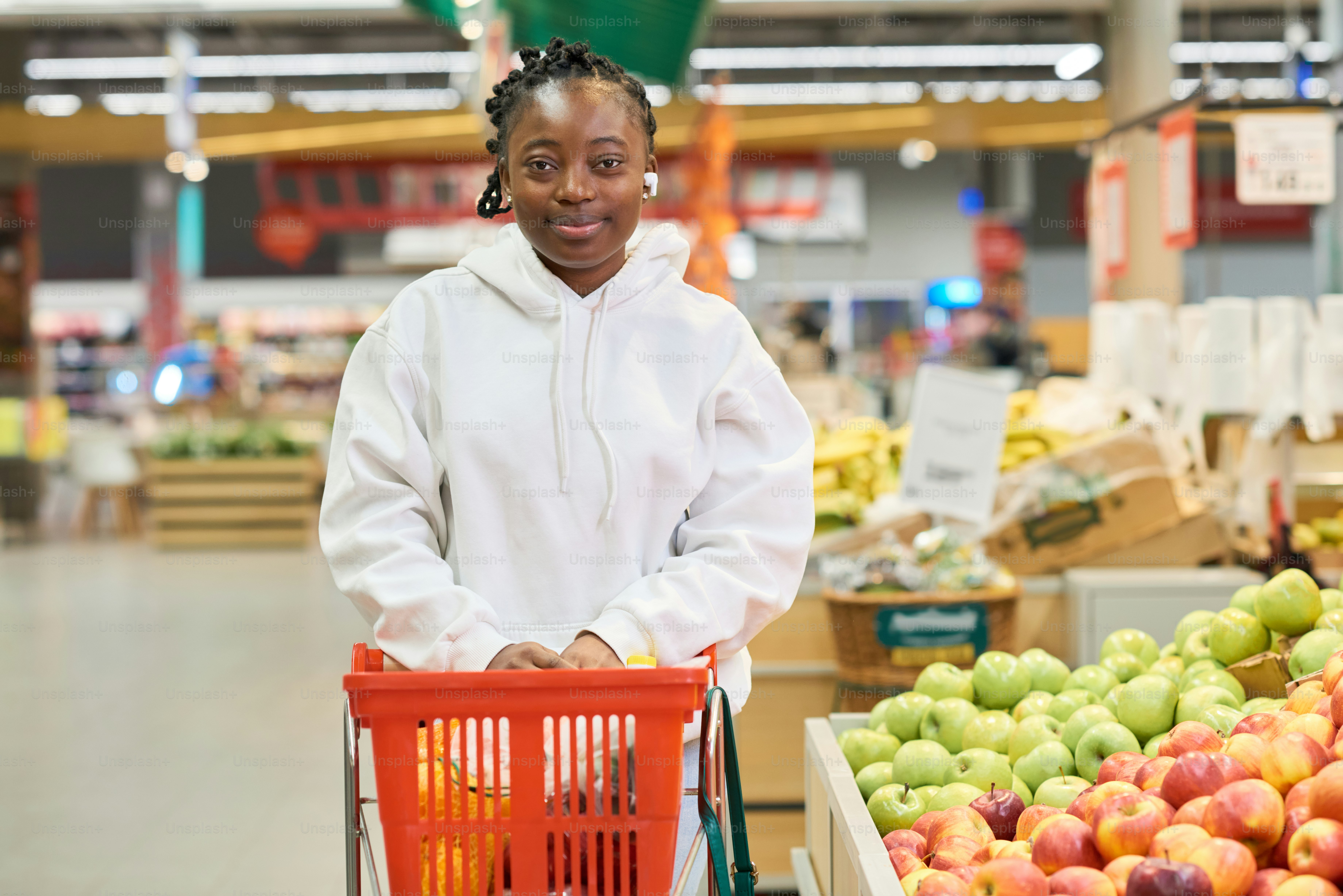 Happy African American girl in white hoodie pushing red shopping cart while moving along display with fresh fruits in grocery department