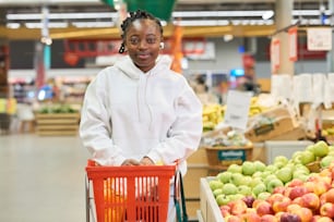 Happy African American girl in white hoodie pushing red shopping cart while moving along display with fresh fruits in grocery department
