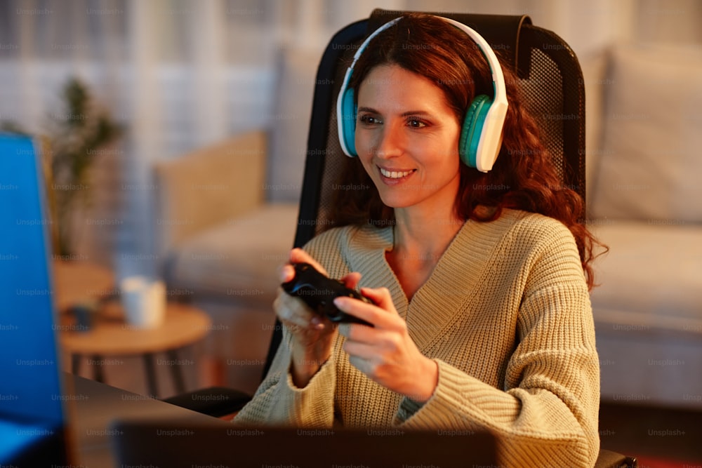 夜遅くにリビングルームでコントローラーを使ってビデオゲームをプレイするヘッドフォンを着た陽気な若い成人白人女性