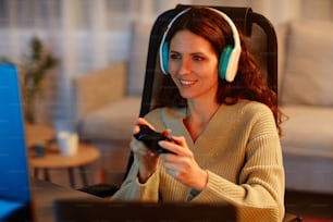 Alegre joven adulta caucásica con auriculares jugando videojuegos usando el controlador en la sala de estar a altas horas de la noche