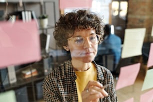 Jovem empresária com cabelos cacheados em óculos fazendo anotações em adesivos pendurados em parede de vidro no escritório