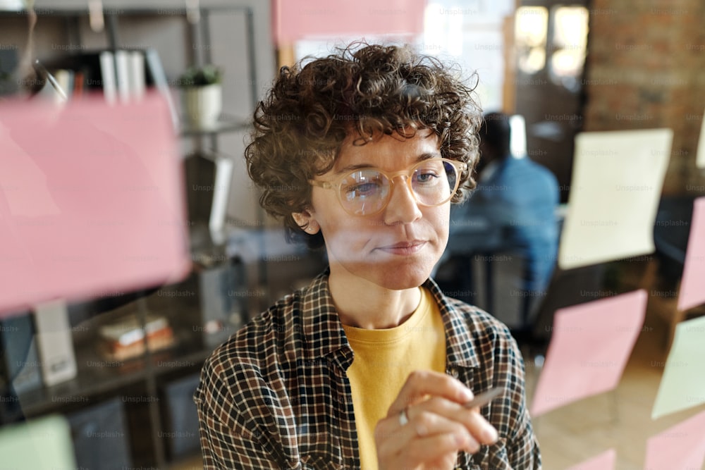 Young businesswoman with curly hair in eyeglasses making notes on stickers hanging on glass wall at office