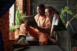 Glückliche junge Dates in Freizeitkleidung, die mit ihren Freunden im Video-Chat kommunizieren, während sie sich am Fenster in der Loft-Wohnung entspannen