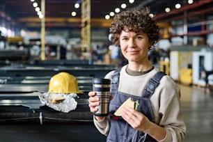 Young pretty female engineer with cup of tea and sandwich looking at camera while having lunch in workshop of industrial factory